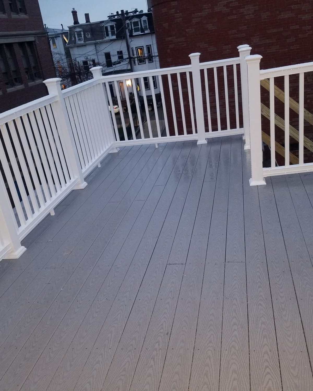 Rooftop deck with gray composite boards and white vinyl railings, brick buildings behind