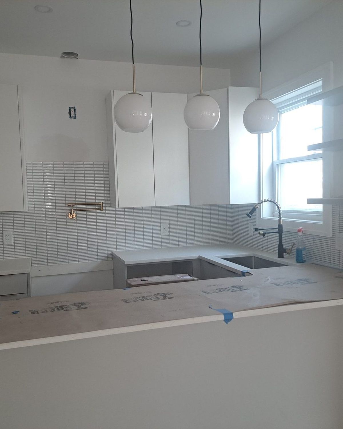 Kitchen under renovation with white cabinets, subway tile backsplash, three pendant lights and countertops covered with protective paper