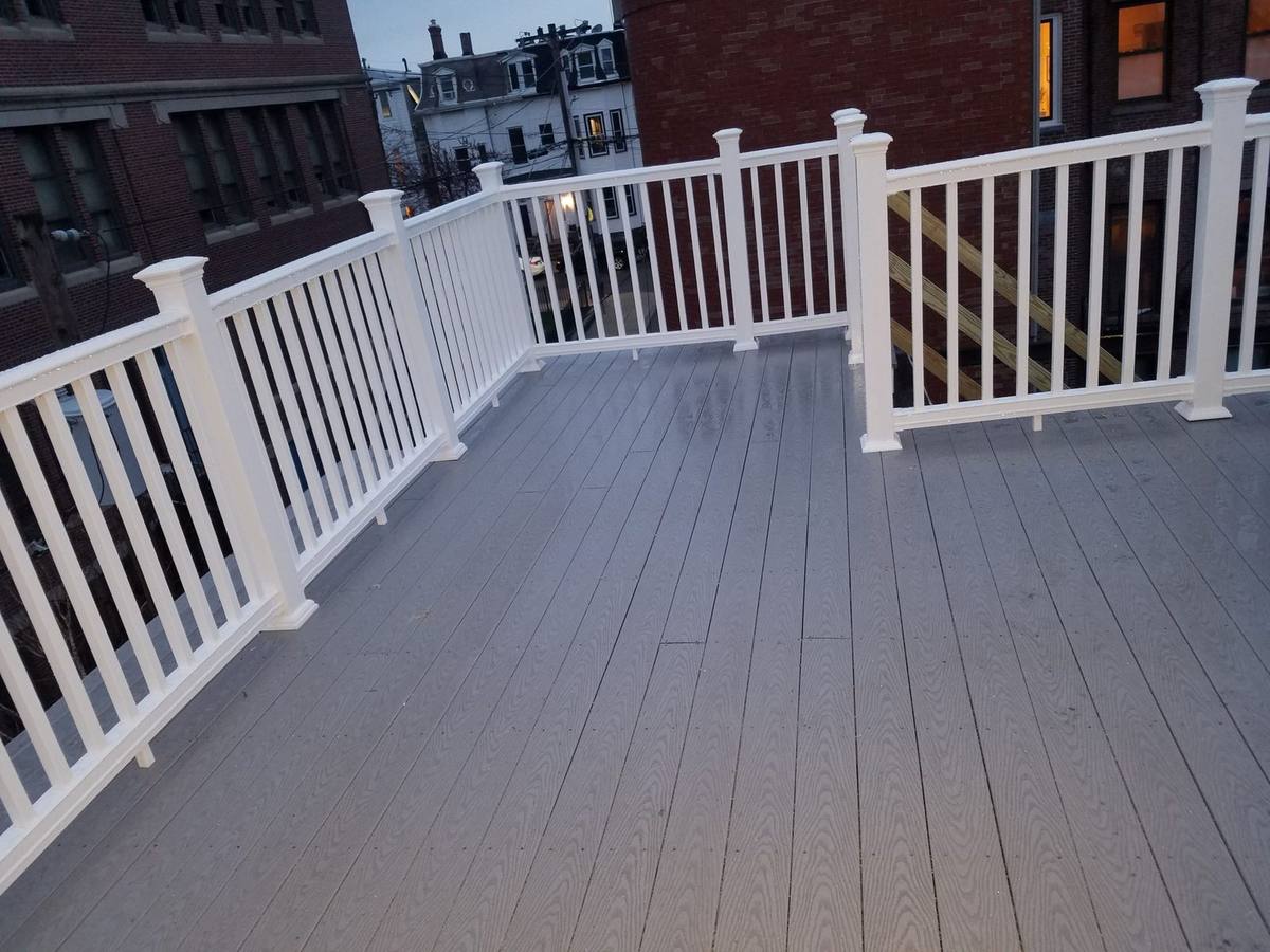 Rooftop deck with gray composite boards and white vinyl railings, brick buildings behind