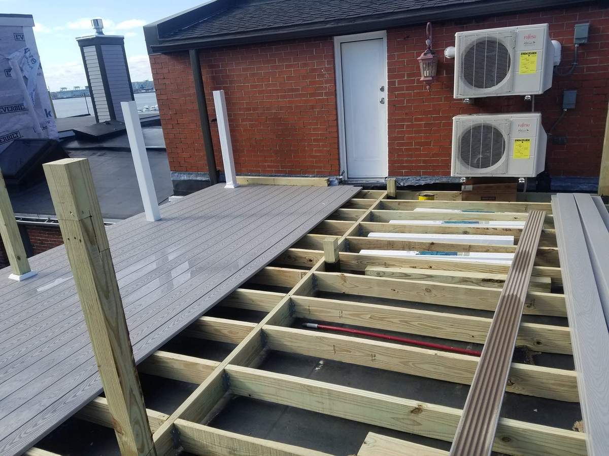 Rooftop deck under construction: composite boards being laid over pressure-treated joists beside a brick wall